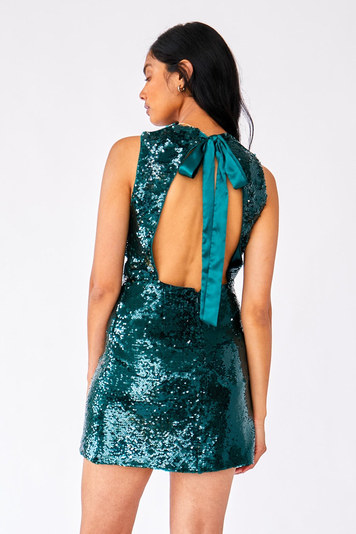 A woman modeling a short, sleeveless, open-back mini dress covered in shimmering teal sequins, shown from the back against a plain light gray studio background.