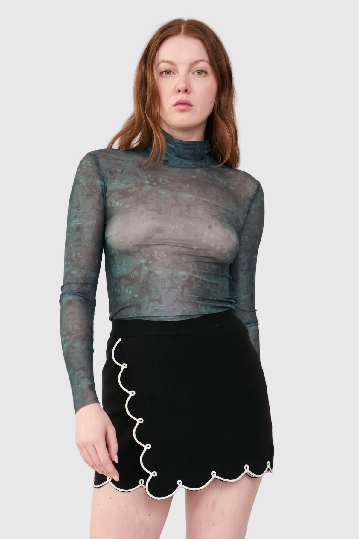 A model wearing a sheer, long-sleeve mock-neck top with a distressed teal and gray pattern, paired with a black skirt featuring a scalloped white trim, against a solid light gray background.