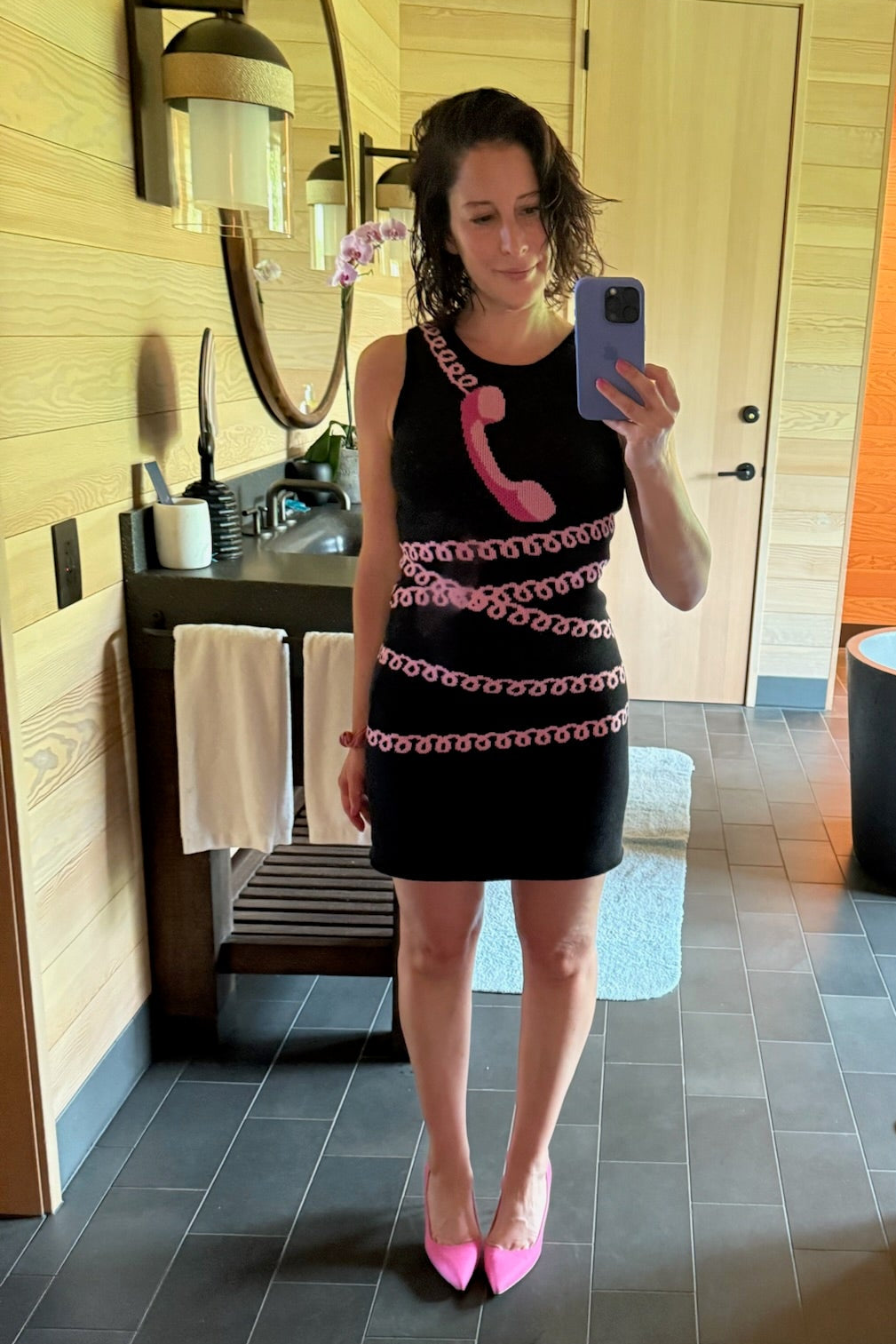A woman takes a mirror selfie in a bathroom wearing a black mini dress with a pink telephone receiver design and pink patterned trim, paired with pink pointed shoes.