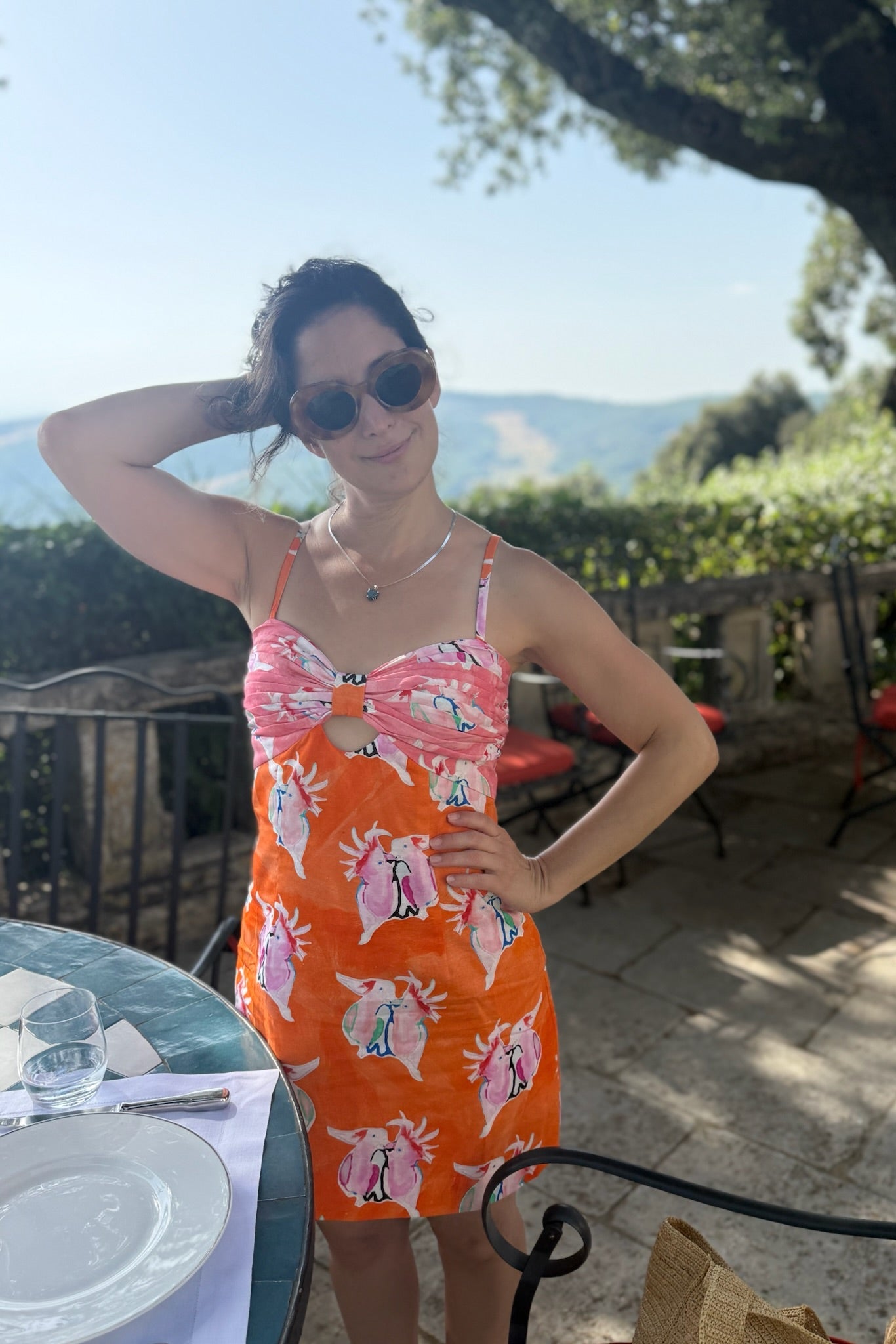 A woman wearing a bright orange and pink bird-print mini dress stands on an outdoor patio with a scenic mountain view in the background.